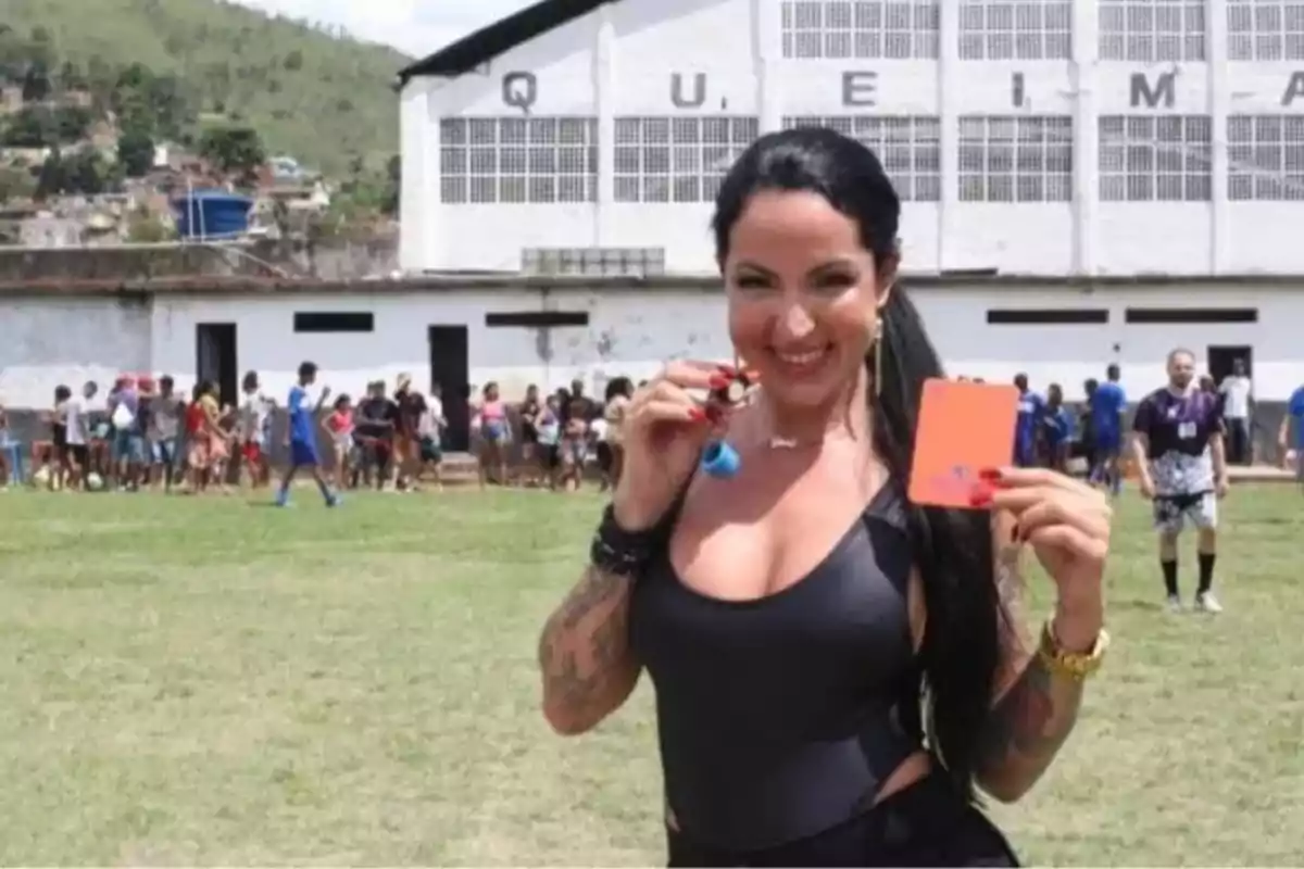 Mujer sonriente sostiene una tarjeta roja y un silbato en un campo de fútbol con personas al fondo Mujer sonriente sostiene una tarjeta roja y un silbato en un campo de fútbol con personas al fondo
