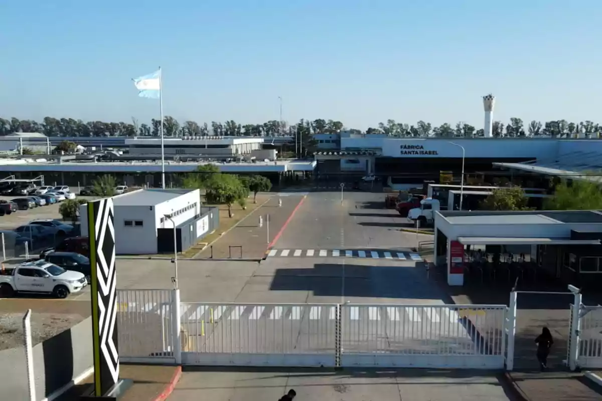 Vista de la entrada principal de la fábrica Santa Isabel con una bandera argentina ondeando y varios autos estacionados Vista de la entrada principal de la fábrica Santa Isabel con una bandera argentina ondeando y varios autos estacionados