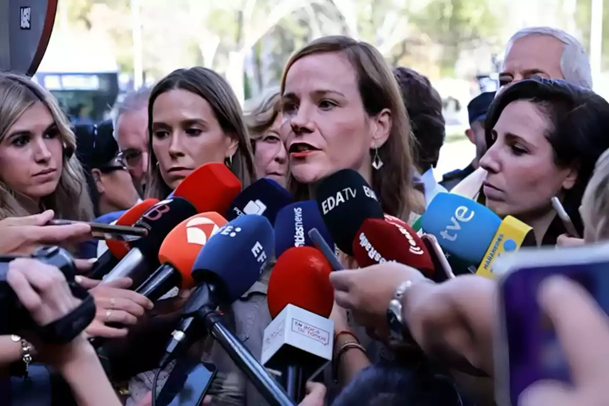 A woman surrounded by journalists answers questions in front of several microphones from different media outlets. A woman surrounded by journalists answers questions in front of several microphones from different media outlets.
