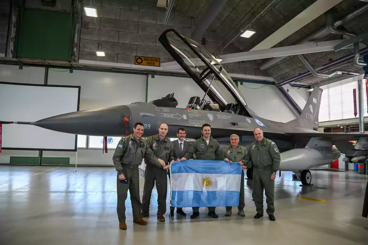 Seis hombres posan sonrientes frente a un avión de combate en un hangar mientras sostienen una bandera de Argentina