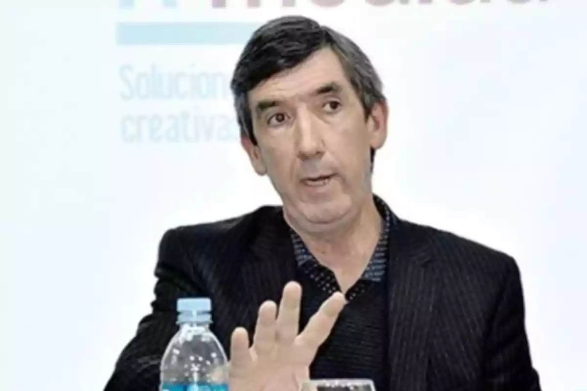Hombre de cabello corto y canoso con saco oscuro hablando y gesticulando con la mano frente a una botella de agua y un vaso Hombre de cabello corto y canoso con saco oscuro hablando y gesticulando con la mano frente a una botella de agua y un vaso