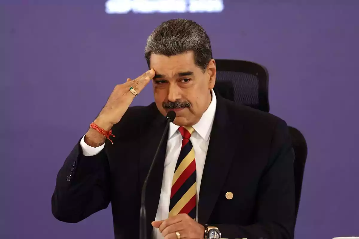 Gray-haired man with a mustache wearing a dark suit and striped tie, sitting in front of a microphone and making a saluting gesture with his hand on his forehead Gray-haired man with a mustache wearing a dark suit and striped tie, sitting in front of a microphone and making a saluting gesture with his hand on his forehead