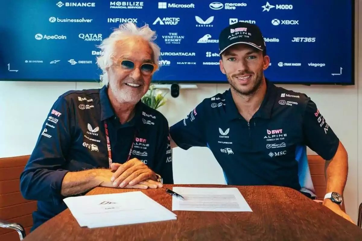 Two men wearing Alpine Formula 1 team clothing smile while sitting at a table with documents and a pen; in the background, a screen with sponsor logos can be seen. Two men wearing Alpine Formula 1 team clothing smile while sitting at a table with documents and a pen; in the background, a screen with sponsor logos can be seen.
