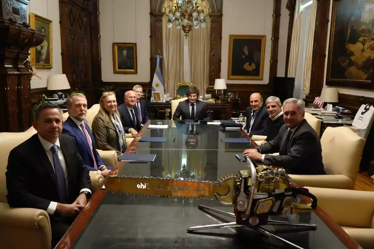 Un grupo de personas con ropa formal sentadas alrededor de una mesa de vidrio en una sala elegante con cuadros en las paredes y una motosierra decorativa sobre la mesa