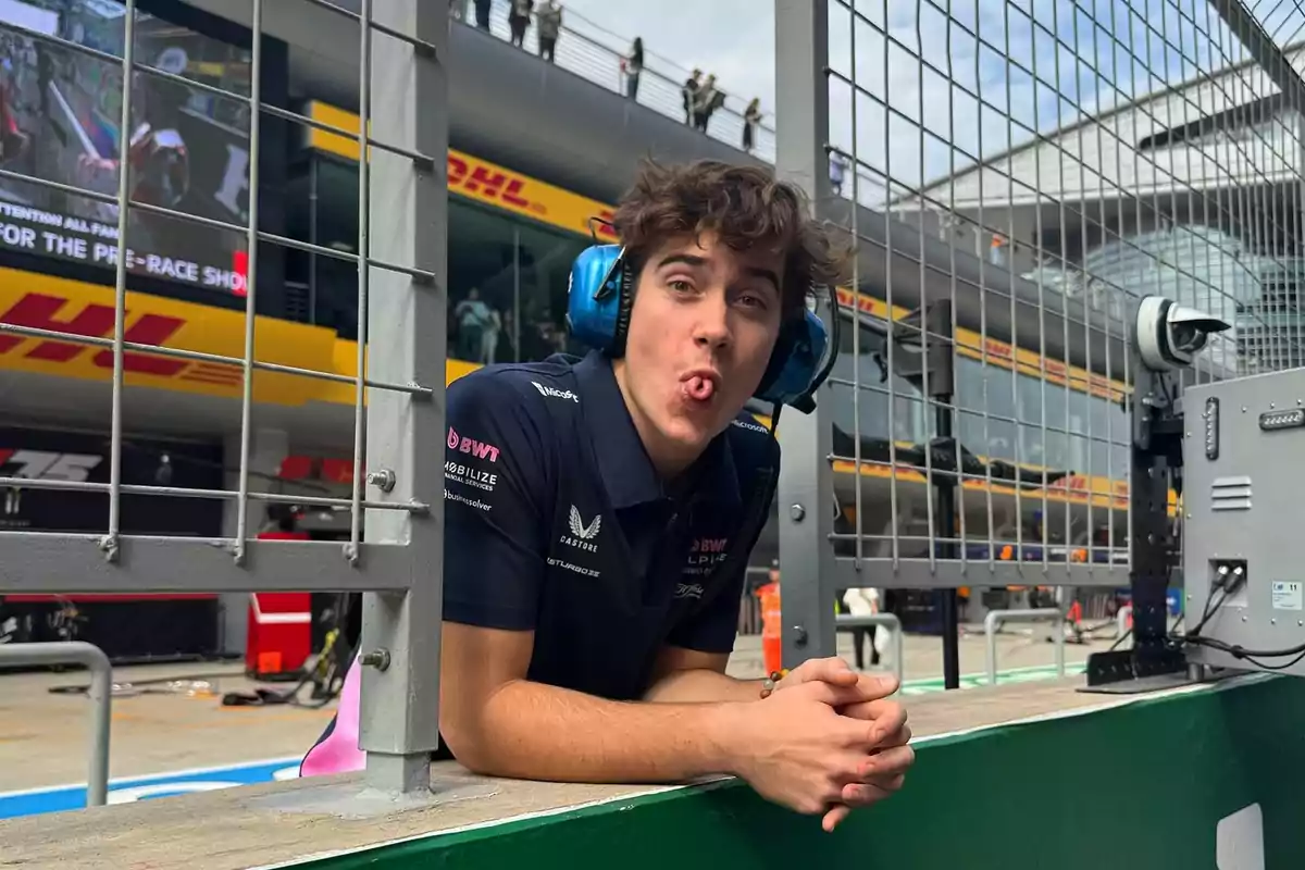 Young man with blue headphones and a racing team jersey making a funny face behind a fence at a motorsport circuit Young man with blue headphones and a racing team jersey making a funny face behind a fence at a motorsport circuit