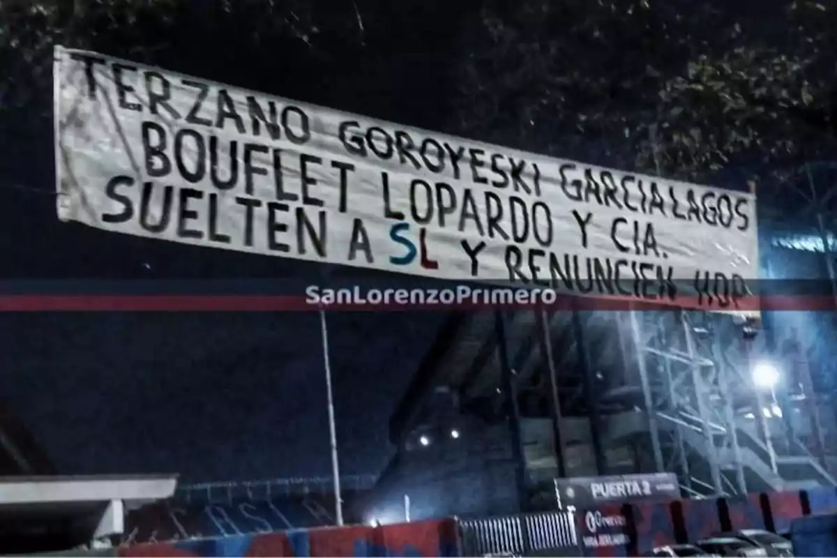 Pancarta colgada en el exterior de un estadio de fútbol durante la noche con nombres y un mensaje dirigido a directivos pidiendo que dejen el club y renuncien
