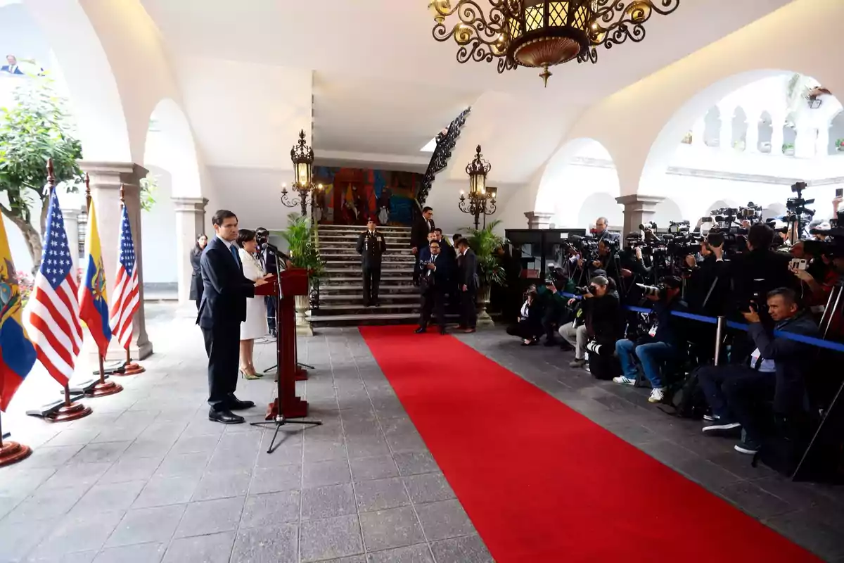 Un funcionario da un discurso en un podio junto a banderas de Ecuador y Estados Unidos mientras numerosos fotógrafos y periodistas capturan el evento en un elegante salón con alfombra roja y lámparas colgantes Un funcionario da un discurso en un podio junto a banderas de Ecuador y Estados Unidos mientras numerosos fotógrafos y periodistas capturan el evento en un elegante salón con alfombra roja y lámparas colgantes