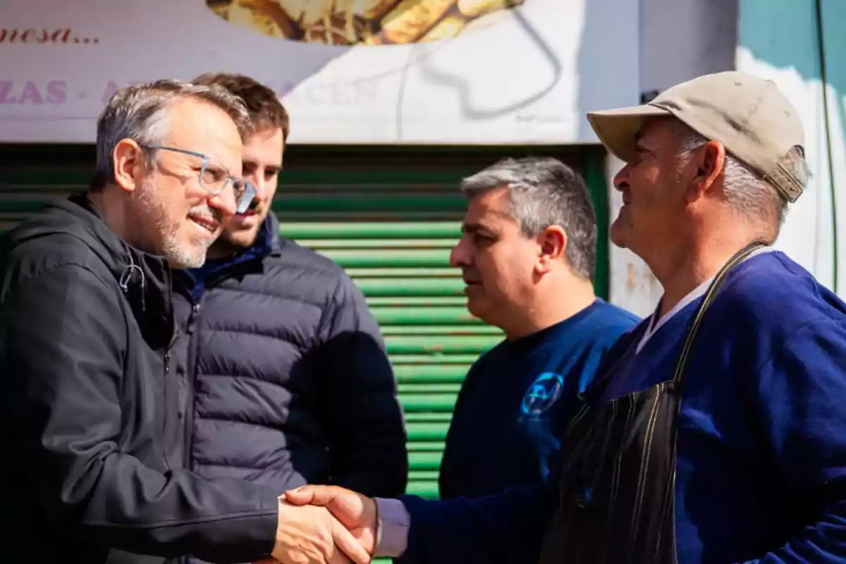 Four men talk and greet each other with a handshake on the street in front of a green shutter. Four men talk and greet each other with a handshake on the street in front of a green shutter.