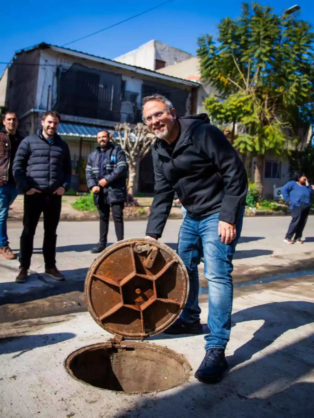 A smiling man opens a manhole cover on a street while other people watch in the background on a sunny day. A smiling man opens a manhole cover on a street while other people watch in the background on a sunny day.