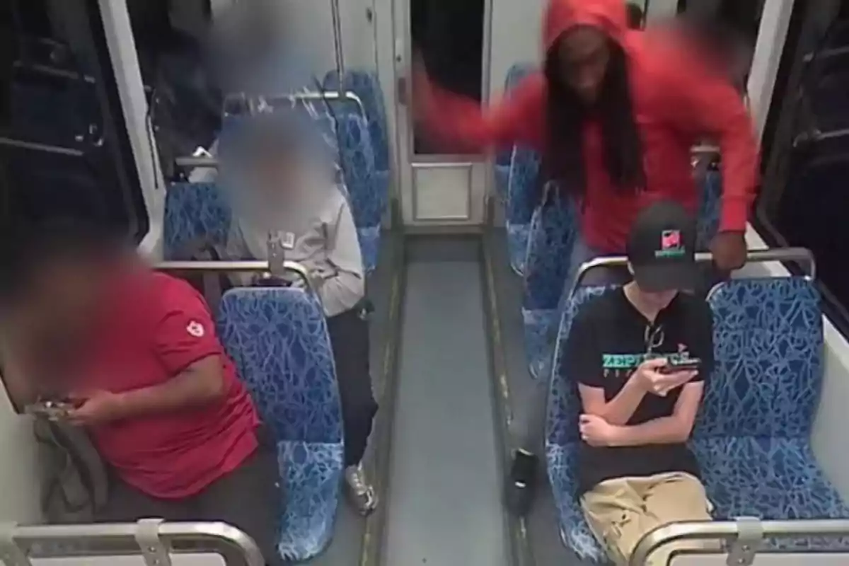 A person in a red hoodie quickly approaches a young man sitting on public transportation while other passengers are distracted using their phones.