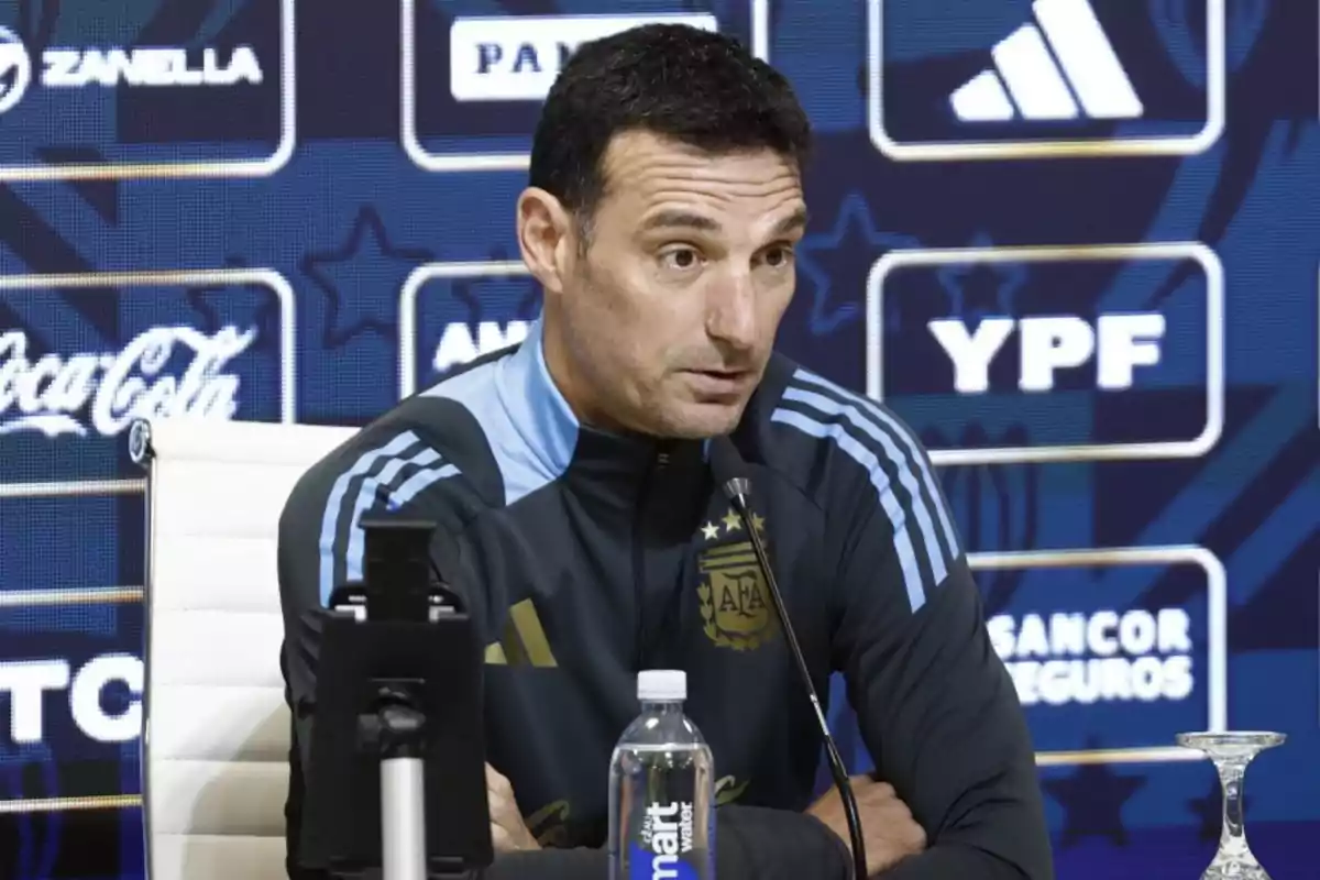 Coach of the Argentine national soccer team speaking at a press conference with sponsor logos in the background and a microphone in front of him
