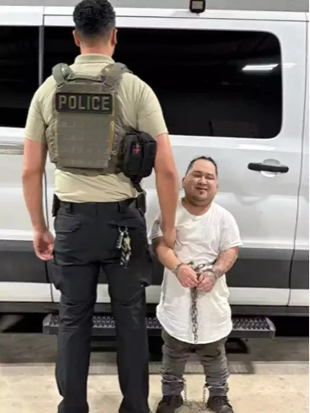 A police officer with his back turned guards a handcuffed and chained man in front of a white van. A police officer with his back turned guards a handcuffed and chained man in front of a white van.