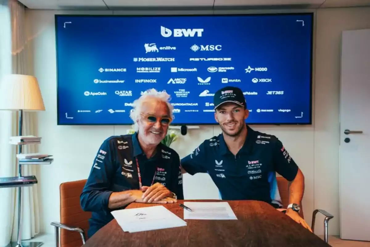 Two men in motorsport team clothing smile while sitting at a table with documents and a pen, with a blue screen displaying sponsor logos in the background. Two men in motorsport team clothing smile while sitting at a table with documents and a pen, with a blue screen displaying sponsor logos in the background.