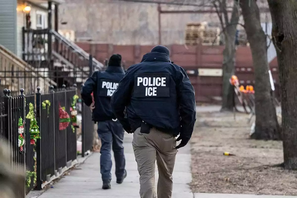 Dos agentes de policía con chaquetas que dicen Police ICE caminan por una acera en un vecindario residencial Dos agentes de policía con chaquetas que dicen Police ICE caminan por una acera en un vecindario residencial