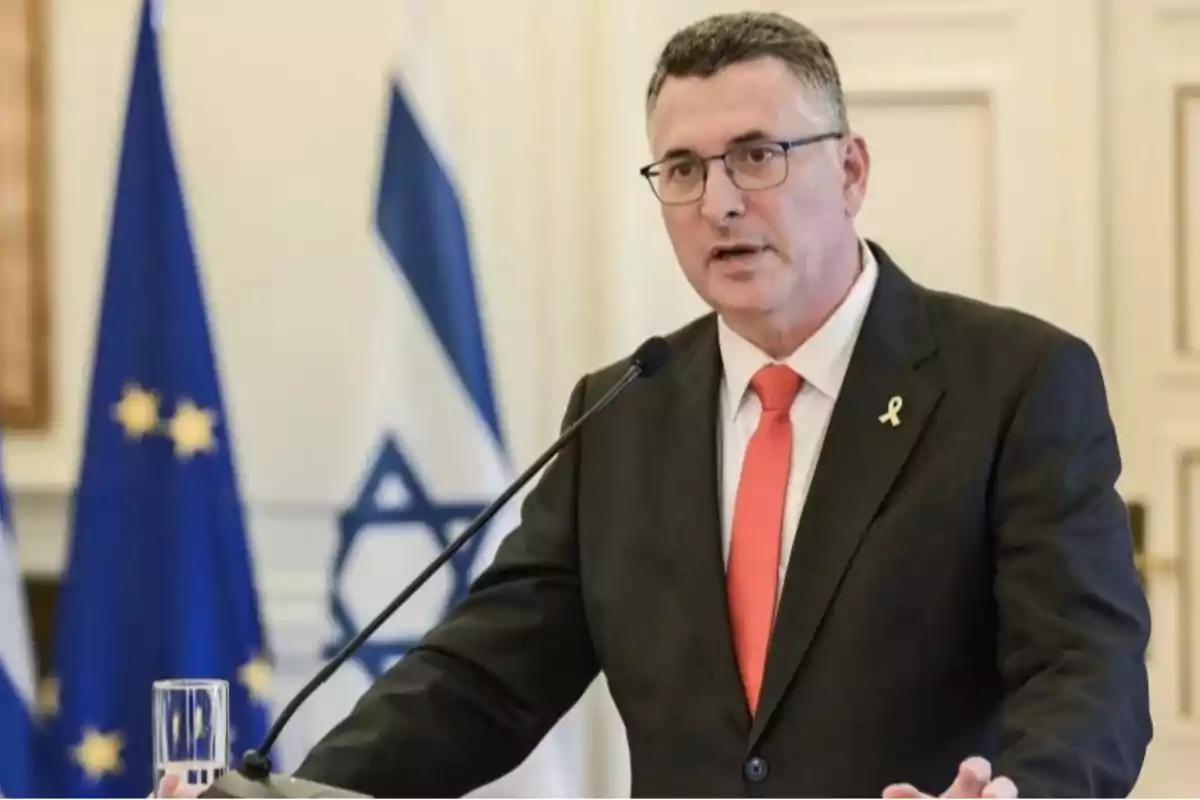 Hombre con gafas y corbata roja hablando en un podio con banderas de la Unión Europea e Israel al fondo