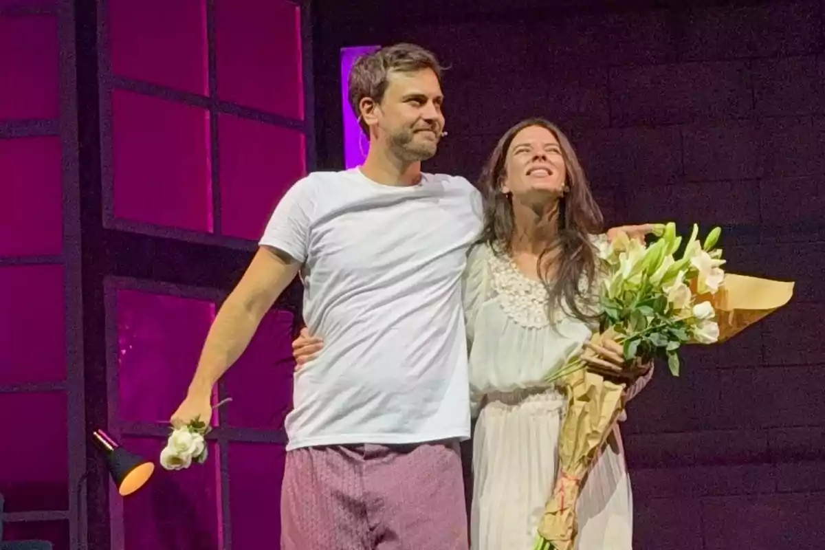 Two people on stage are hugging; one is holding a bouquet of flowers and the other a lamp and a flower. Both are smiling under a violet light in the background.