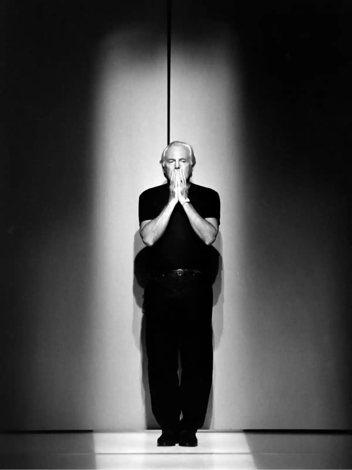 Gray-haired man dressed in black standing at the center of a stage with his hands together in front of his face under a light that shines on him Gray-haired man dressed in black standing at the center of a stage with his hands together in front of his face under a light that shines on him