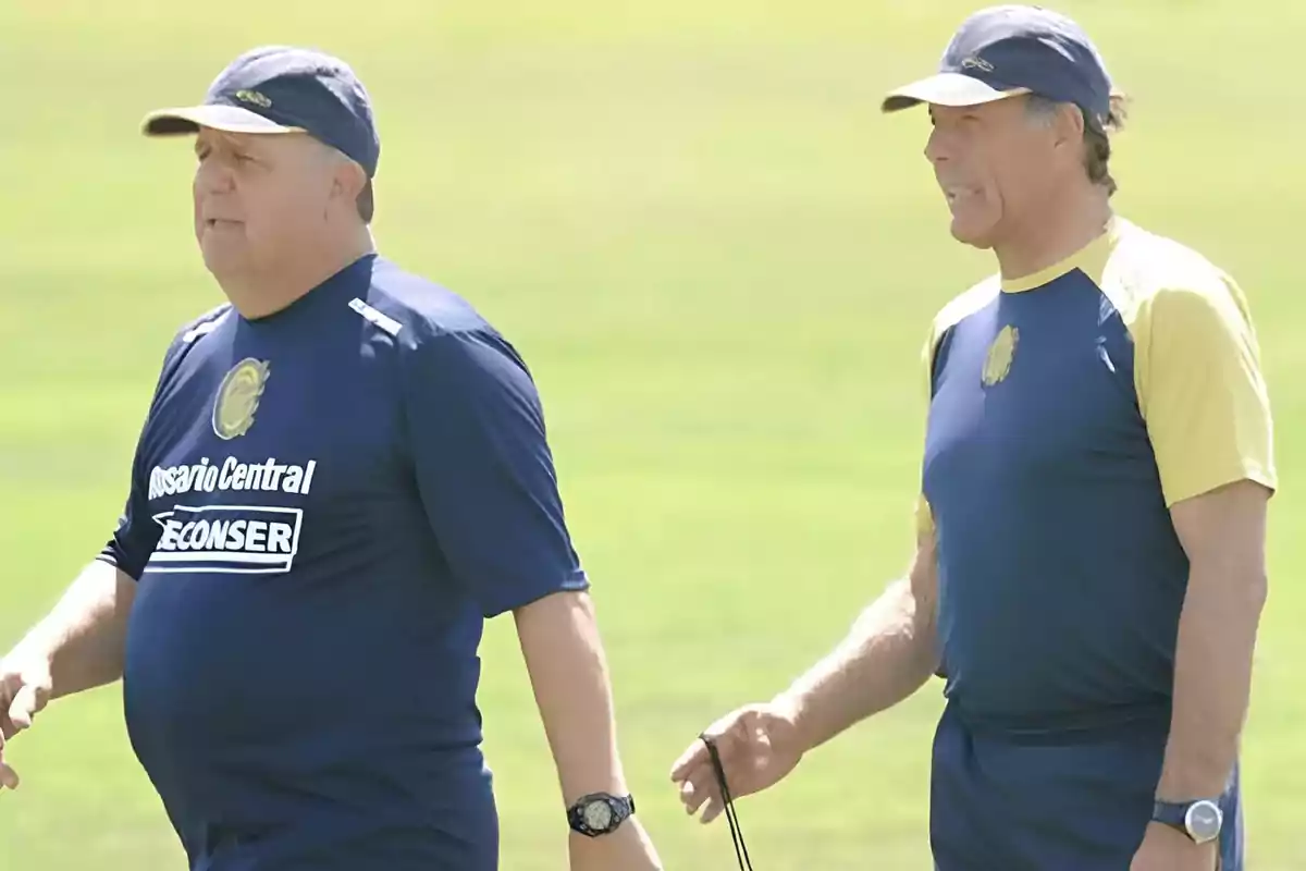 Two men wearing Rosario Central sportswear walk across a grassy field during the day. Two men wearing Rosario Central sportswear walk across a grassy field during the day.