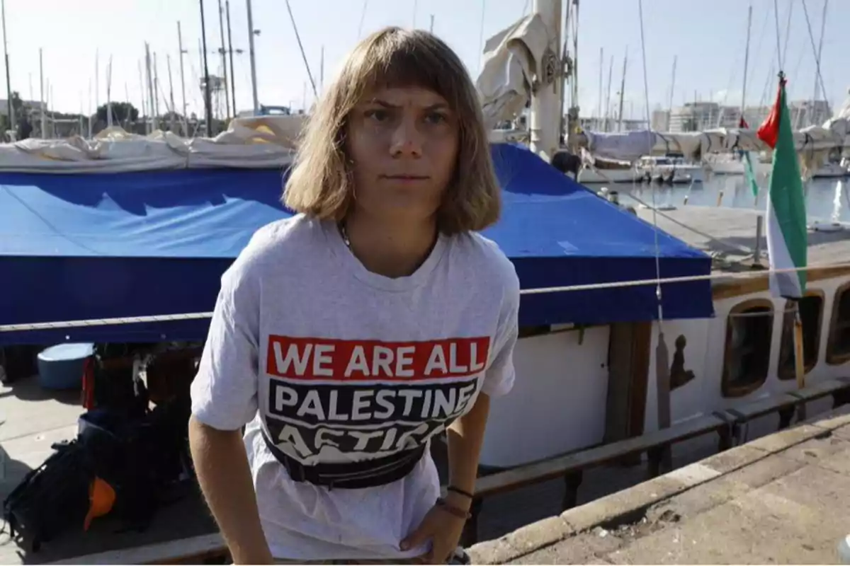 Persona con cabello corto y camiseta con mensaje de apoyo a Palestina de pie frente a un barco en un puerto