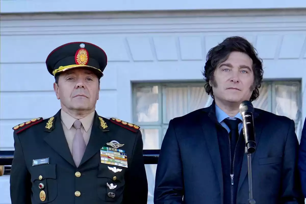 A military officer in dress uniform and a man in a dark suit are standing side by side in front of a white building, the man in the suit is holding a microphone. A military officer in dress uniform and a man in a dark suit are standing side by side in front of a white building, the man in the suit is holding a microphone.