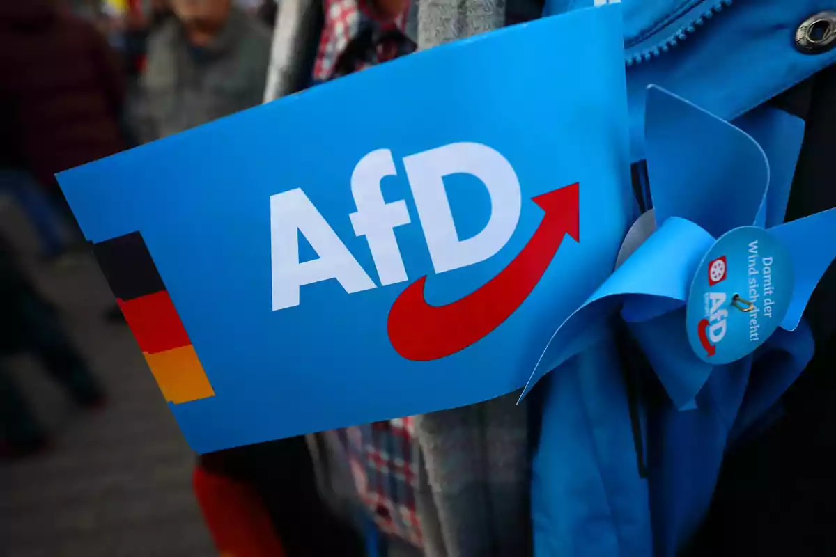 Bandera azul con el logo de AfD y una flecha roja, junto a un molinillo de papel azul y una pequeña bandera de Alemania en la esquina Bandera azul con el logo de AfD y una flecha roja, junto a un molinillo de papel azul y una pequeña bandera de Alemania en la esquina