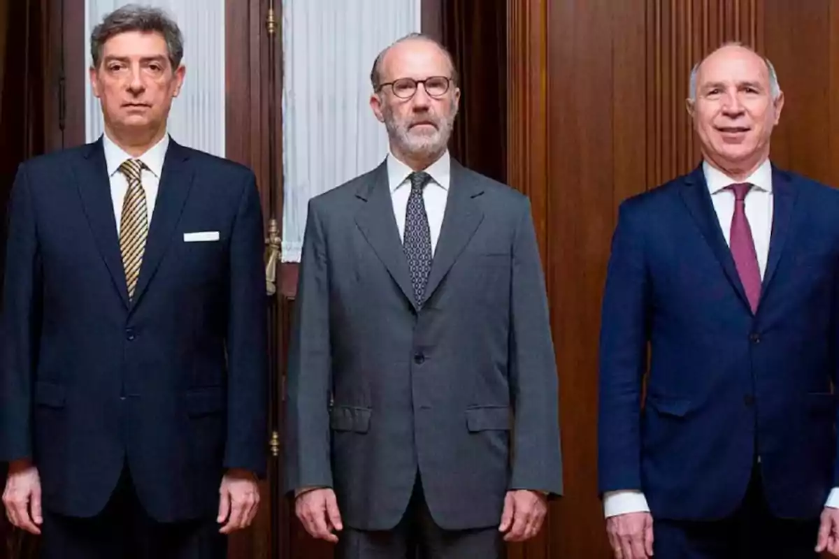 Tres hombres de traje posan de pie en un ambiente formal con paredes de madera y cortinas blancas al fondo Tres hombres de traje posan de pie en un ambiente formal con paredes de madera y cortinas blancas al fondo