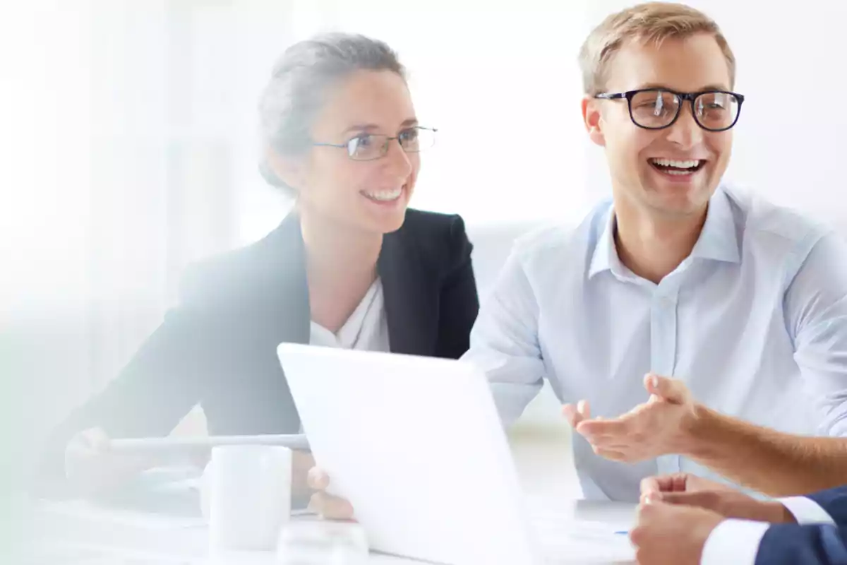 Dos personas sonrientes con gafas conversan en una reunión de trabajo frente a una laptop Dos personas sonrientes con gafas conversan en una reunión de trabajo frente a una laptop
