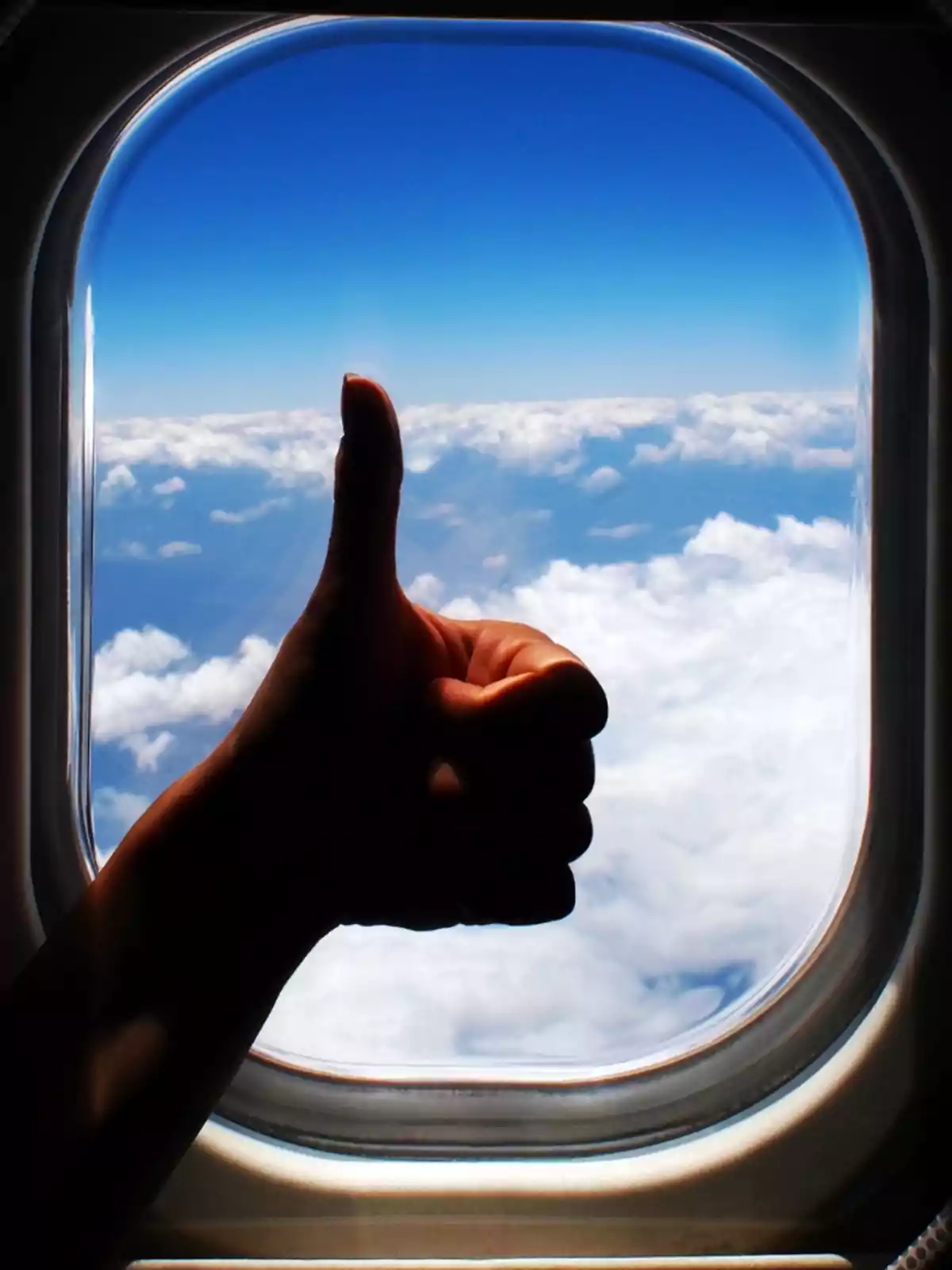 Mano haciendo señal de pulgar arriba frente a la ventana de un avión con nubes y cielo azul de fondo Mano haciendo señal de pulgar arriba frente a la ventana de un avión con nubes y cielo azul de fondo