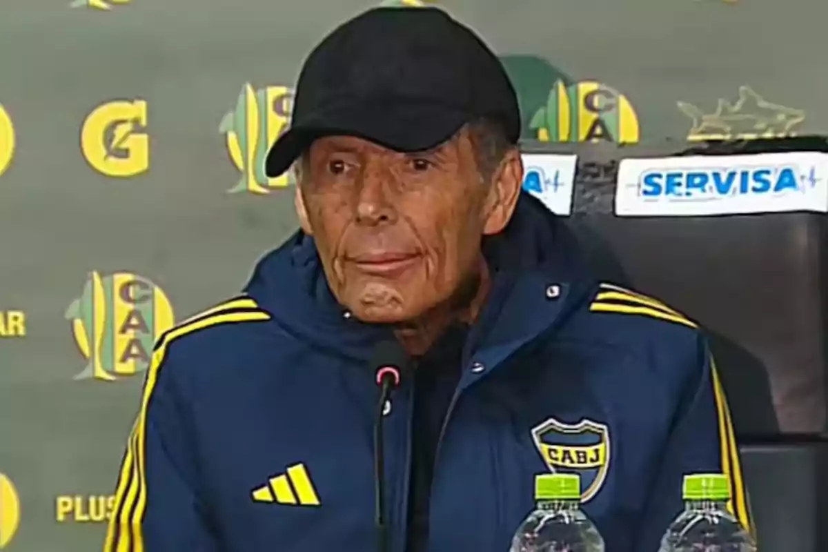 Older man wearing a black cap and a blue Boca Juniors jacket at a press conference