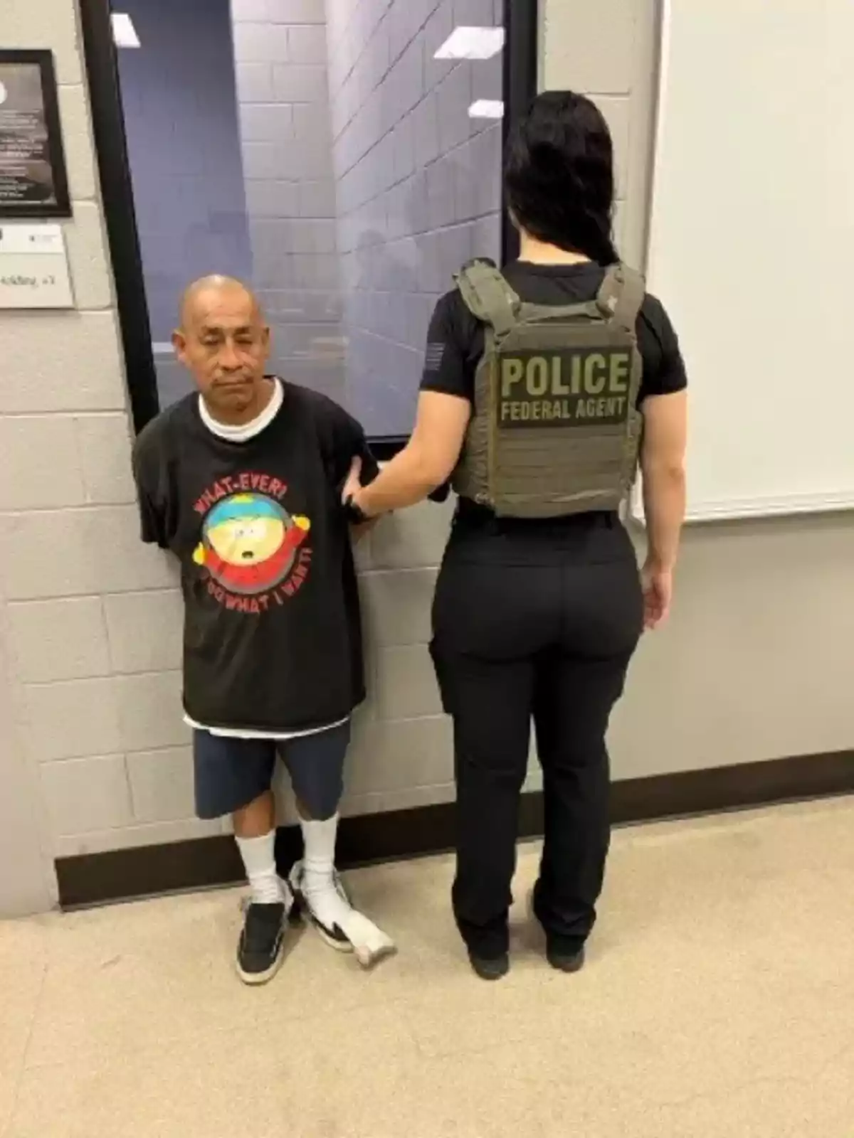 A man wearing a black t-jersey and shorts is standing next to a federal agent wearing a bulletproof vest inside a police station. A man wearing a black t-jersey and shorts is standing next to a federal agent wearing a bulletproof vest inside a police station.