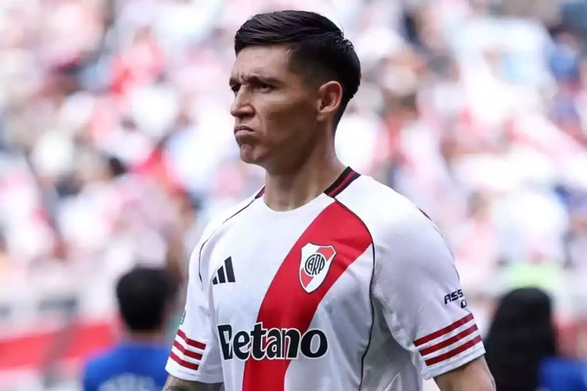 Soccer player with a serious expression wearing River Plate’s white uniform with a red sash in a stadium full of fans.