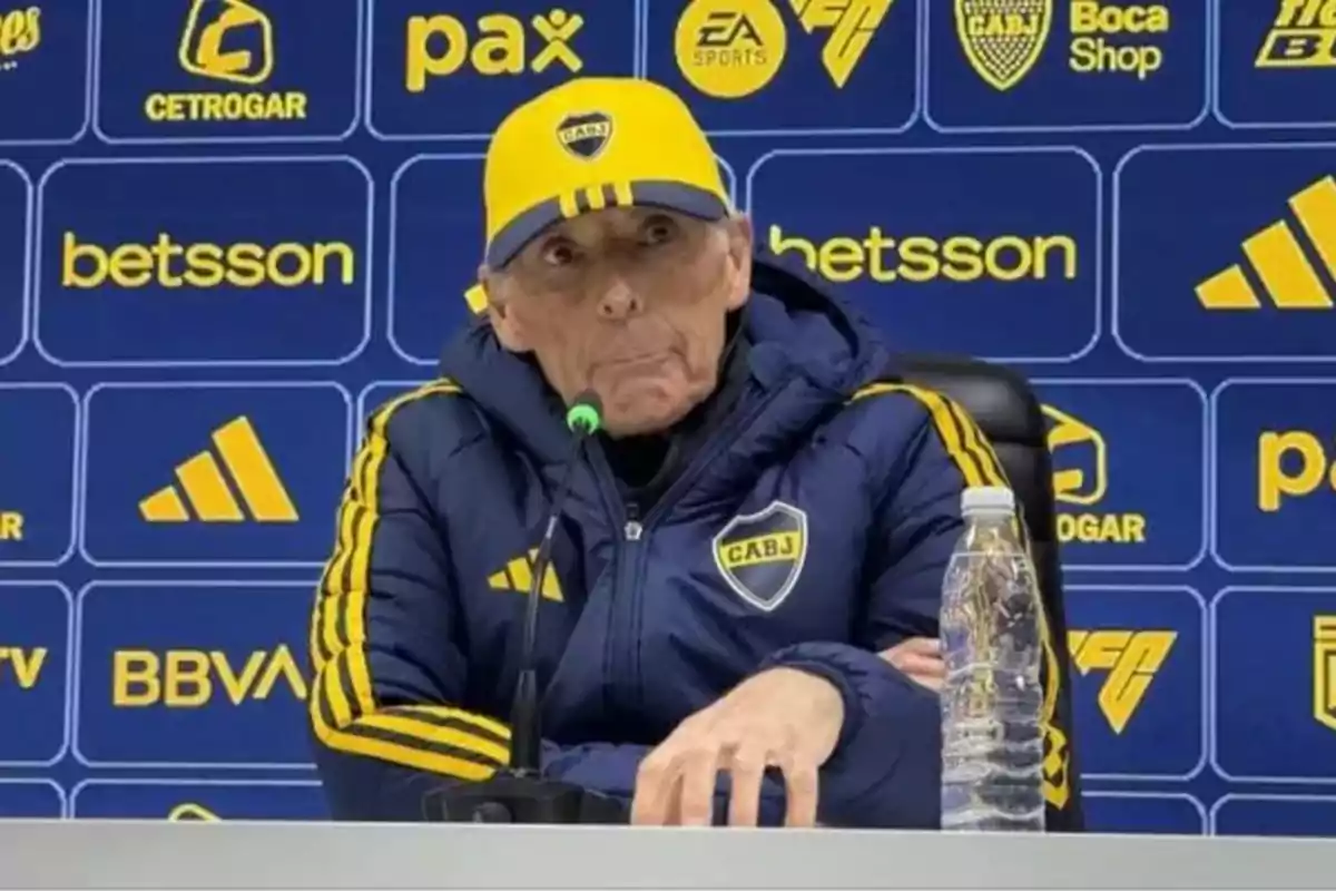 Entrenador de Boca Juniors con gorra y campera del club hablando en una conferencia de prensa frente a un fondo azul con logos de patrocinadores y una botella de agua sobre la mesa