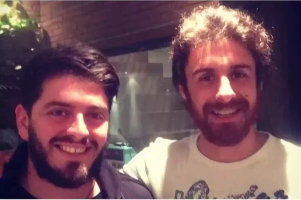 Two young bearded men smile together in a well-lit indoor setting. Two young bearded men smile together in a well-lit indoor setting.