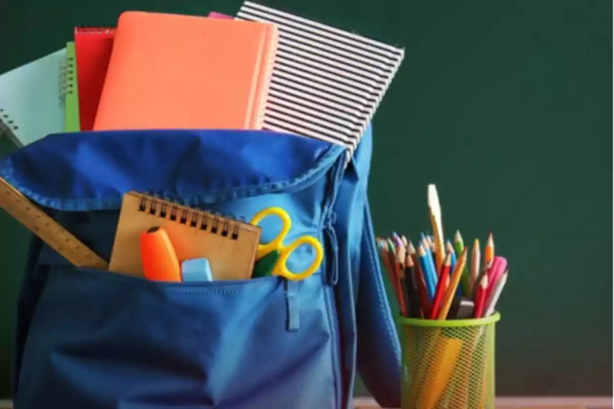 Mochila azul llena de útiles escolares junto a un portalápices verde con colores frente a un pizarrón verde