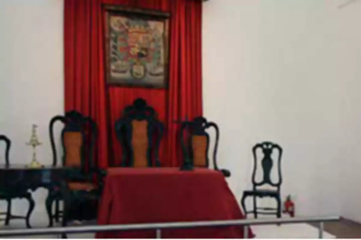 Old room with carved wooden chairs and upholstered backs, table and red curtains, on a white wall with a framed coat of arms Old room with carved wooden chairs and upholstered backs, table and red curtains, on a white wall with a framed coat of arms