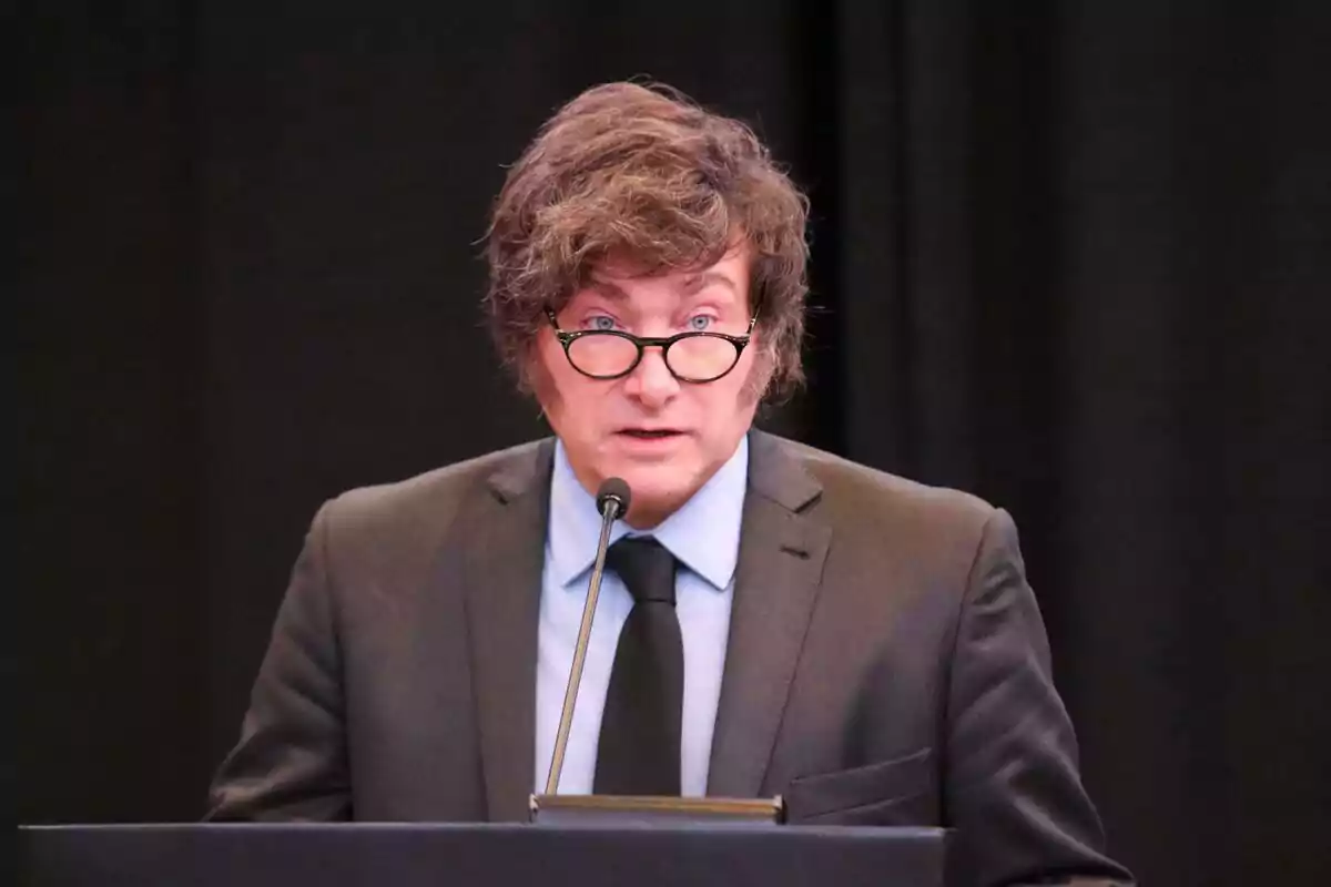 Brown-haired man with glasses speaking in front of a microphone against a black background, wearing a dark suit
