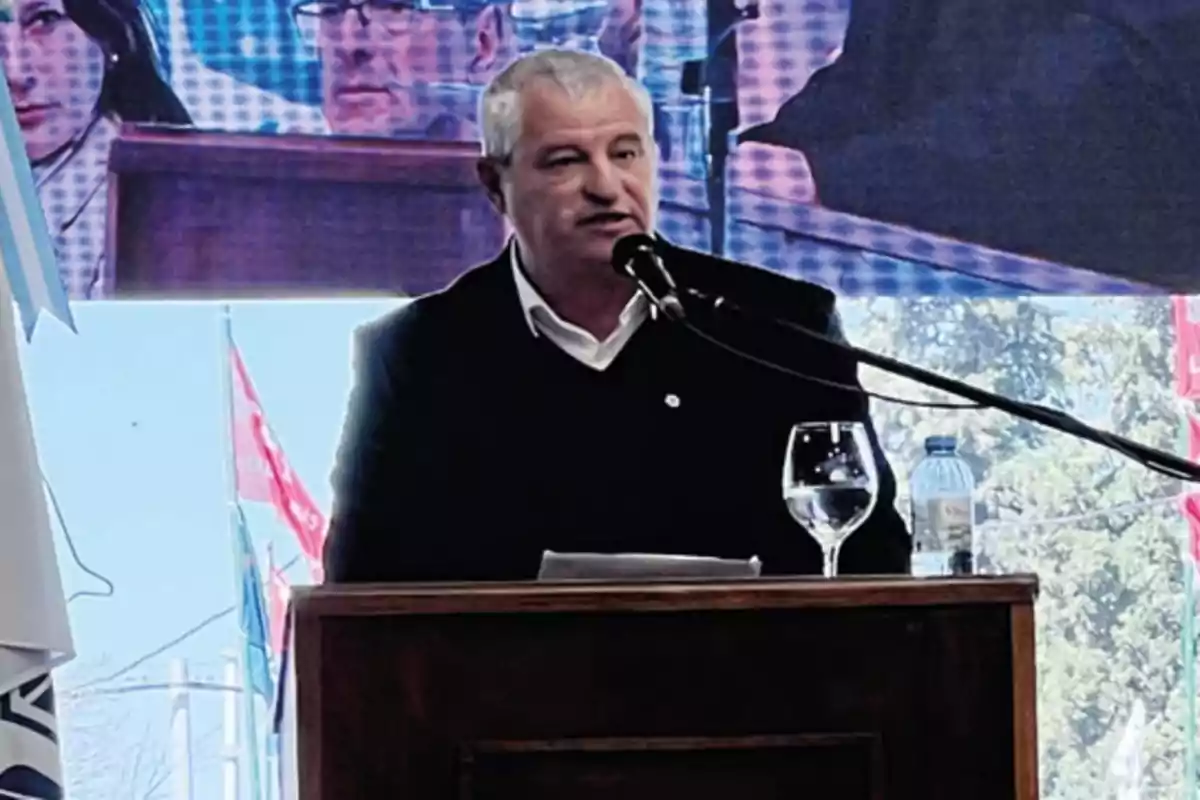 Gray-haired man speaking at a podium with a microphone, water glass and bottle, background with flags and a giant screen.