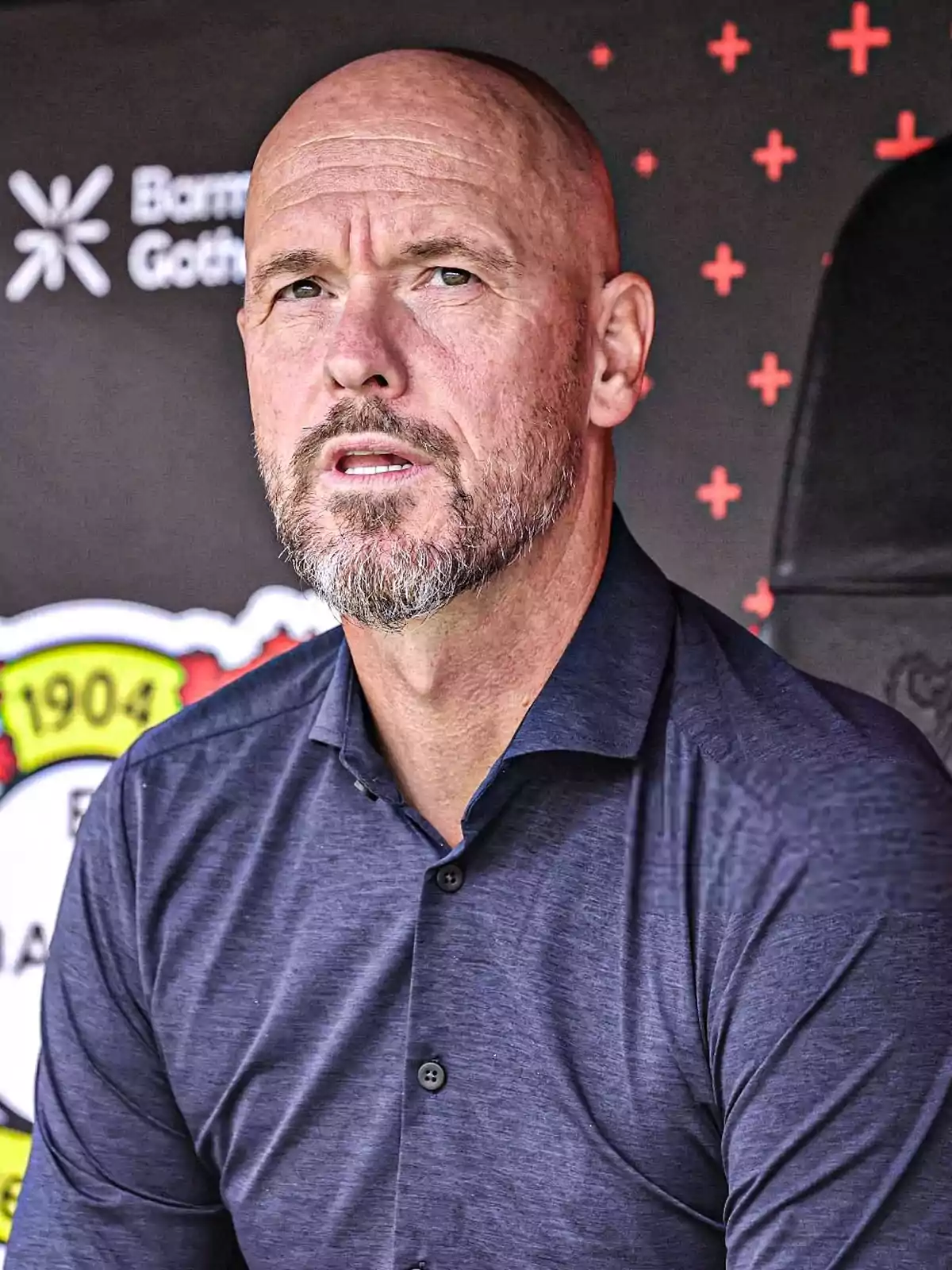 Bald man with a beard and a blue jersey sitting in front of a dark background with red cross symbols and part of a logo visible on the left. Bald man with a beard and a blue jersey sitting in front of a dark background with red cross symbols and part of a logo visible on the left.