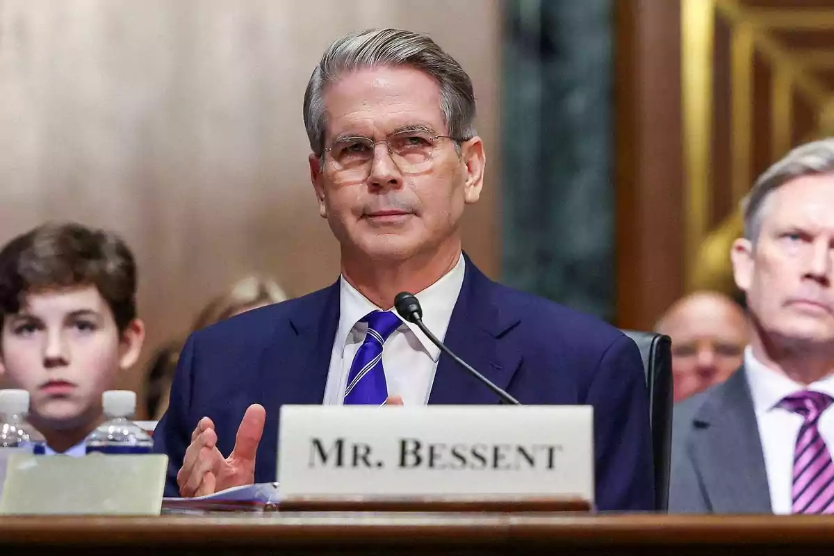 Hombre de cabello canoso y gafas usando traje azul y corbata, sentado frente a un micrófono en una audiencia pública, con un letrero que dice Mr. Bessent y personas al fondo. Hombre de cabello canoso y gafas usando traje azul y corbata, sentado frente a un micrófono en una audiencia pública, con un letrero que dice Mr. Bessent y personas al fondo.