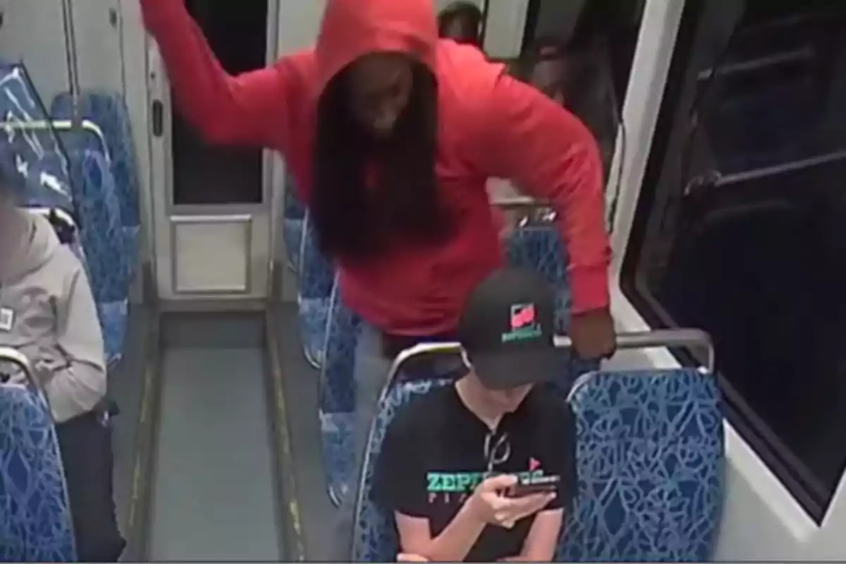Person wearing a red hoodie standing and raising their arm behind another person who is sitting and using their phone on public transportation