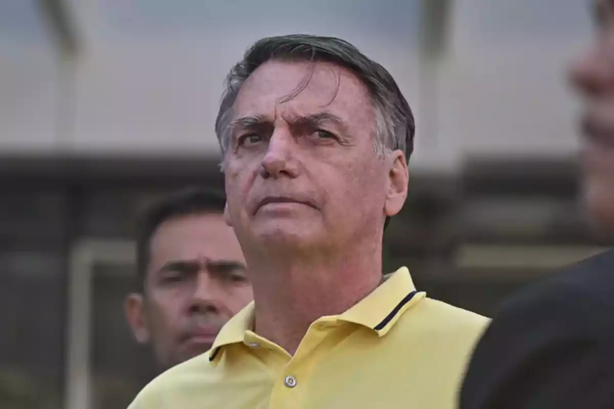 Hombre de cabello canoso y expresión seria viste una camiseta amarilla y está al aire libre con otras personas desenfocadas al fondo Hombre de cabello canoso y expresión seria viste una camiseta amarilla y está al aire libre con otras personas desenfocadas al fondo