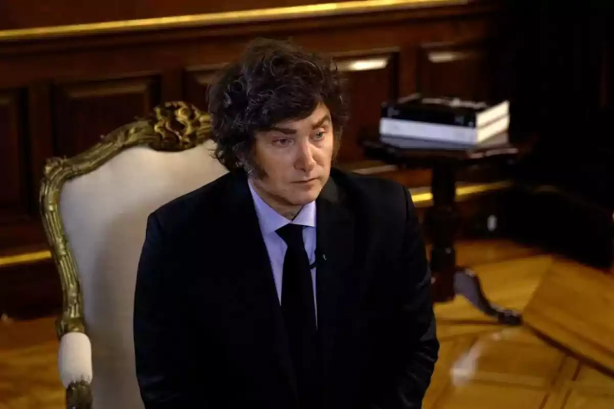 Curly-haired man in a dark suit sitting on an elegant chair in a room with wooden walls and parquet flooring