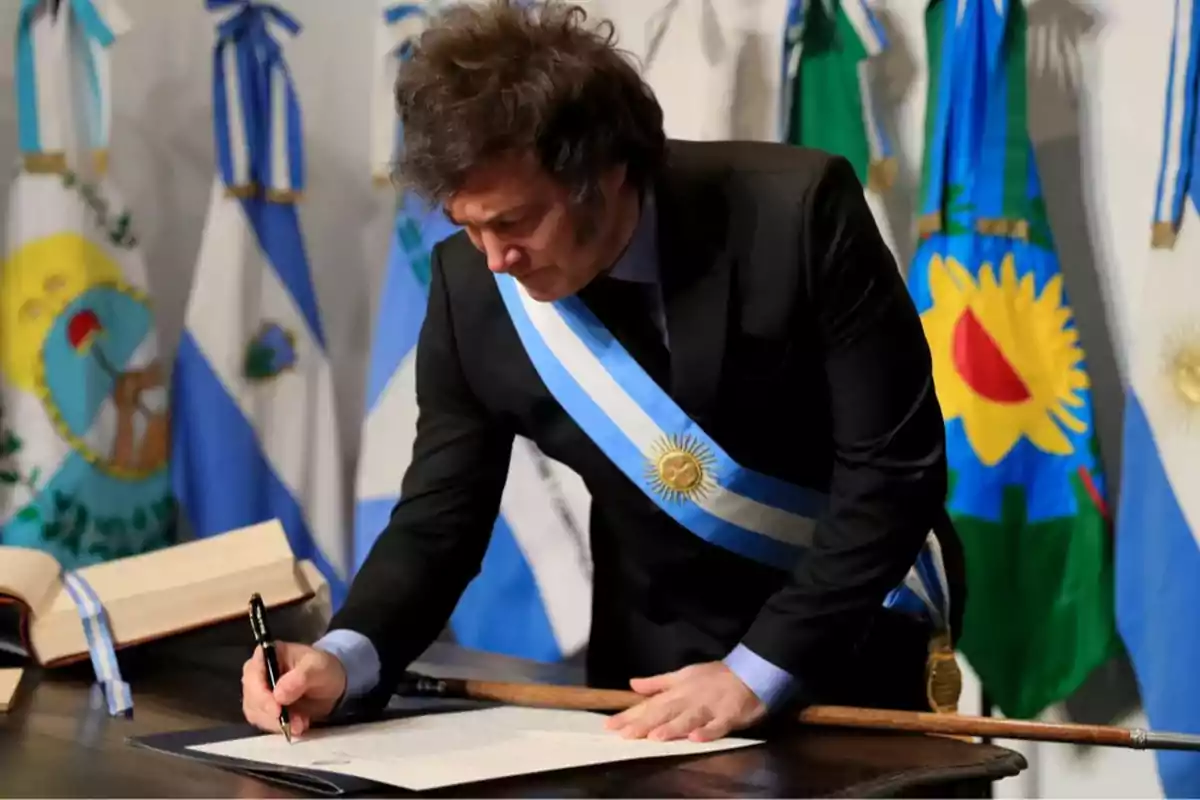 Hombre con banda presidencial argentina firmando un documento oficial en una ceremonia solemne rodeado de banderas.