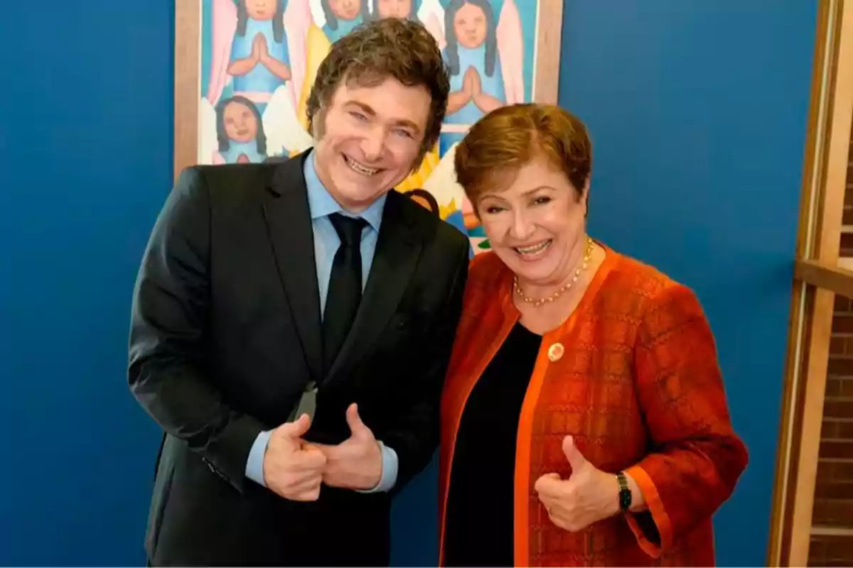 Two people smile and pose together giving a thumbs-up gesture in front of a blue wall and a colorful painting in the background.