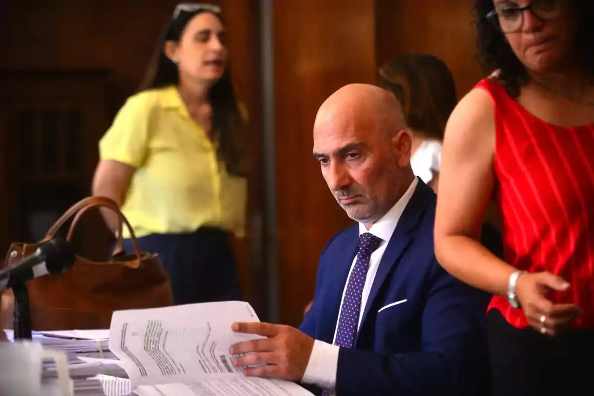 Man in a blue suit reviewing documents at a table while two women talk in the background in an office Man in a blue suit reviewing documents at a table while two women talk in the background in an office