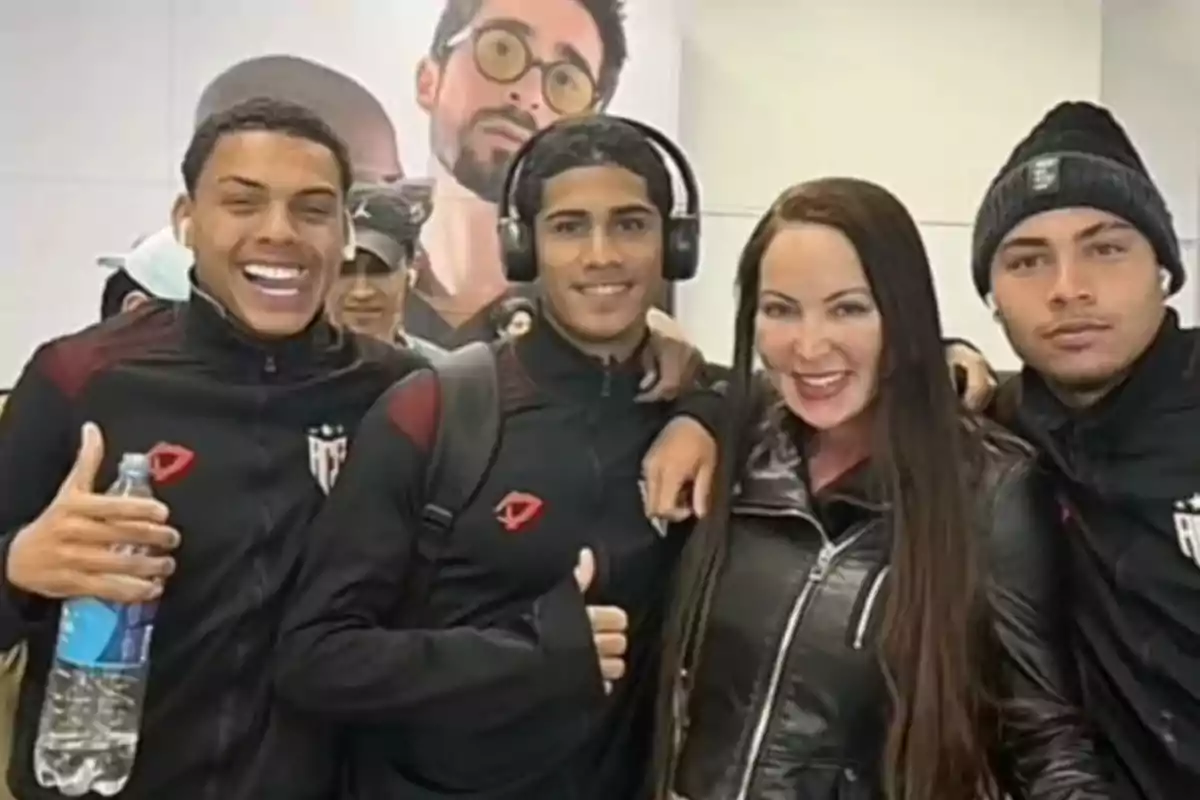 Tres jóvenes con ropa deportiva negra y una mujer con chaqueta de cuero posan sonrientes para la cámara en un ambiente interior