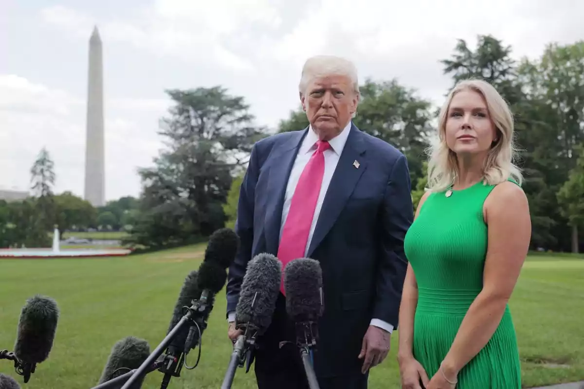 A man in a suit and pink tie and a woman in a green dress pose in front of several microphones in a garden with the Washington Monument in the background. A man in a suit and pink tie and a woman in a green dress pose in front of several microphones in a garden with the Washington Monument in the background.