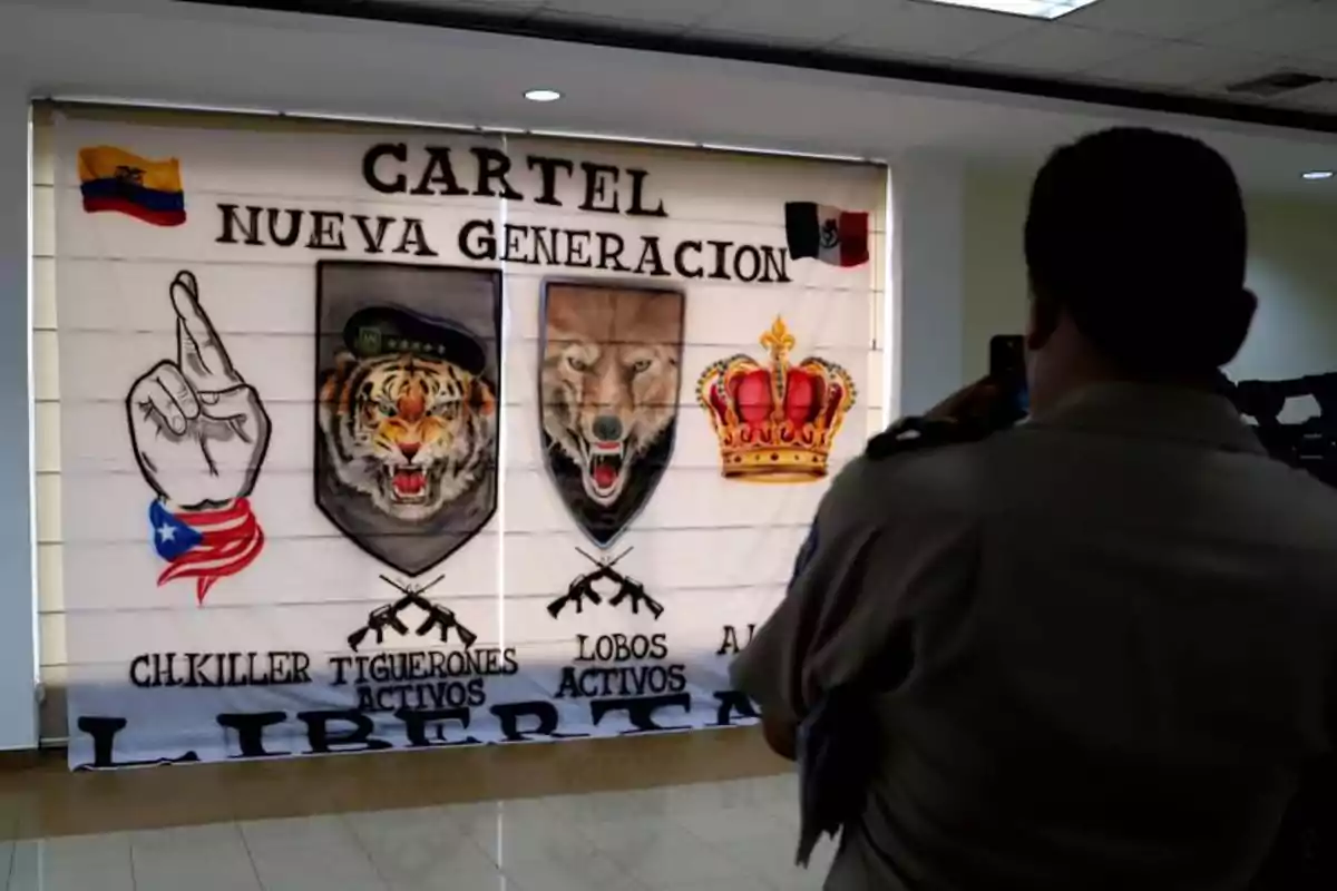 A uniformed man takes a photo of a banner with symbols, crests of a tiger and a wolf, a crown, flags of Ecuador and Mexico, and the text Cartel Nueva Generación. A uniformed man takes a photo of a banner with symbols, crests of a tiger and a wolf, a crown, flags of Ecuador and Mexico, and the text Cartel Nueva Generación.