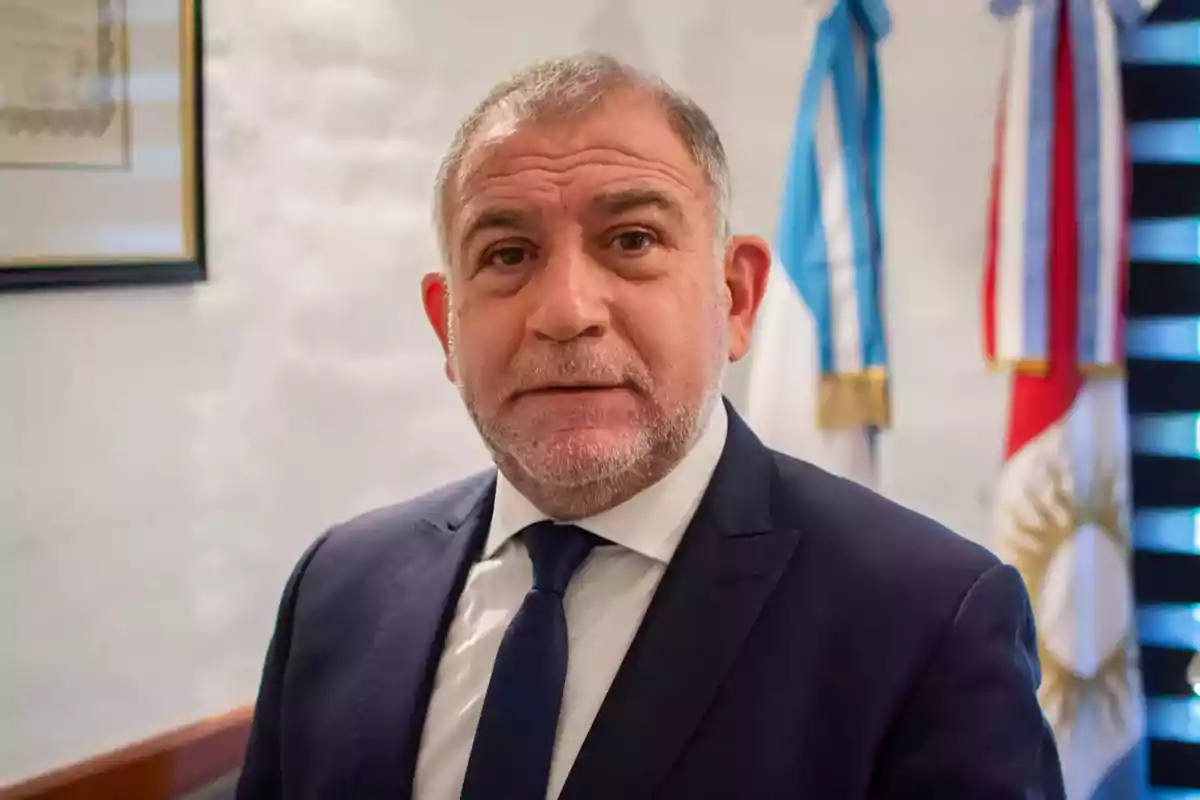 Gray-haired man with a short beard wearing a blue suit and tie, posing in an office with flags in the background.