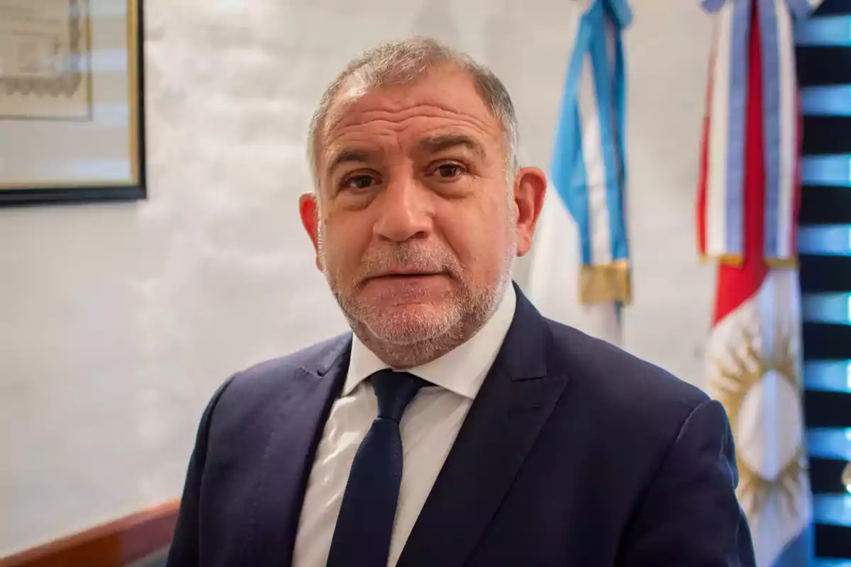 Gray-haired man with a short beard wearing a blue suit and tie, standing in an office with Argentine flags in the background Gray-haired man with a short beard wearing a blue suit and tie, standing in an office with Argentine flags in the background